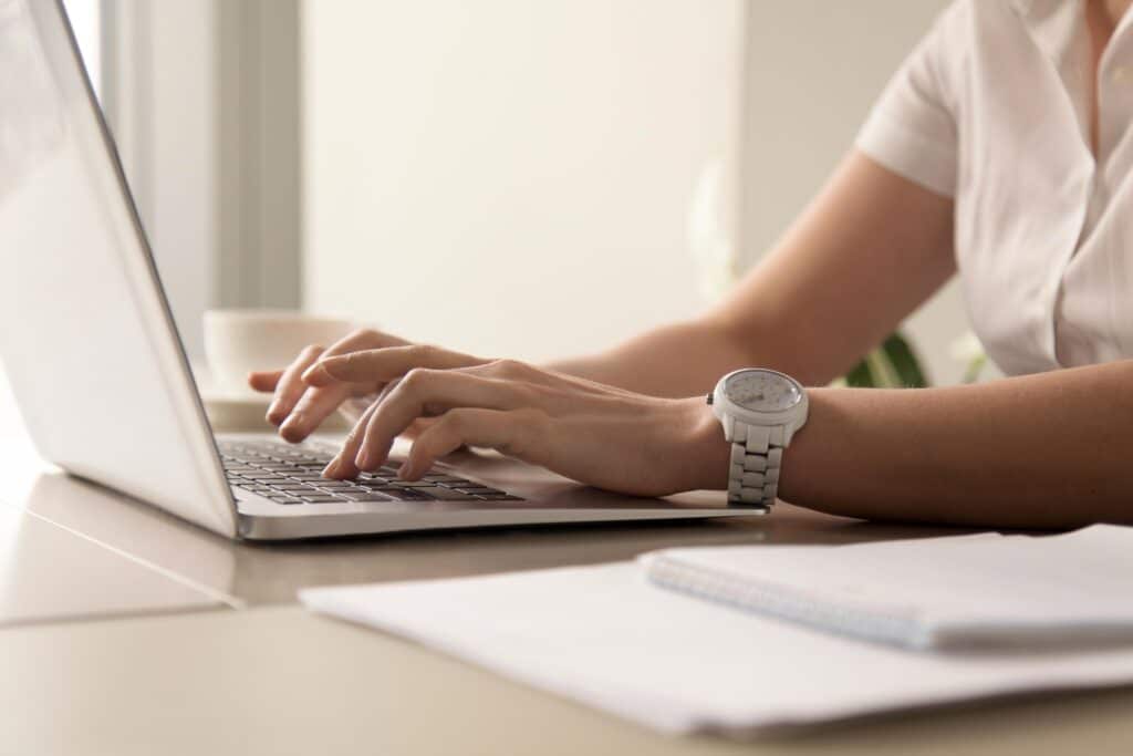 Krupni plan ruku osobe u bijeloj košulji koja tipka na laptopu, s bijelim satom na ruci. U pozadini se vidi šalica kafe i radni stol. Slika simbolizira produktivan rad u coworking prostoru.