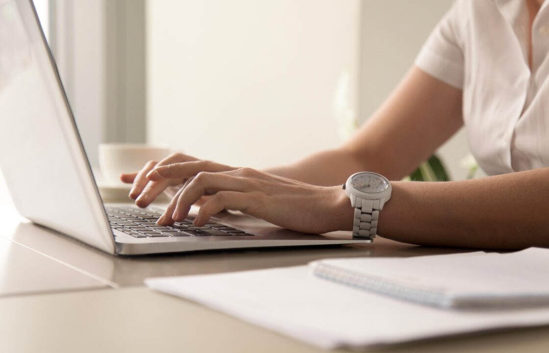 Krupni plan ruku osobe u bijeloj košulji koja tipka na laptopu, s bijelim satom na ruci. U pozadini se vidi šalica kafe i radni stol. Slika simbolizira produktivan rad u coworking prostoru.